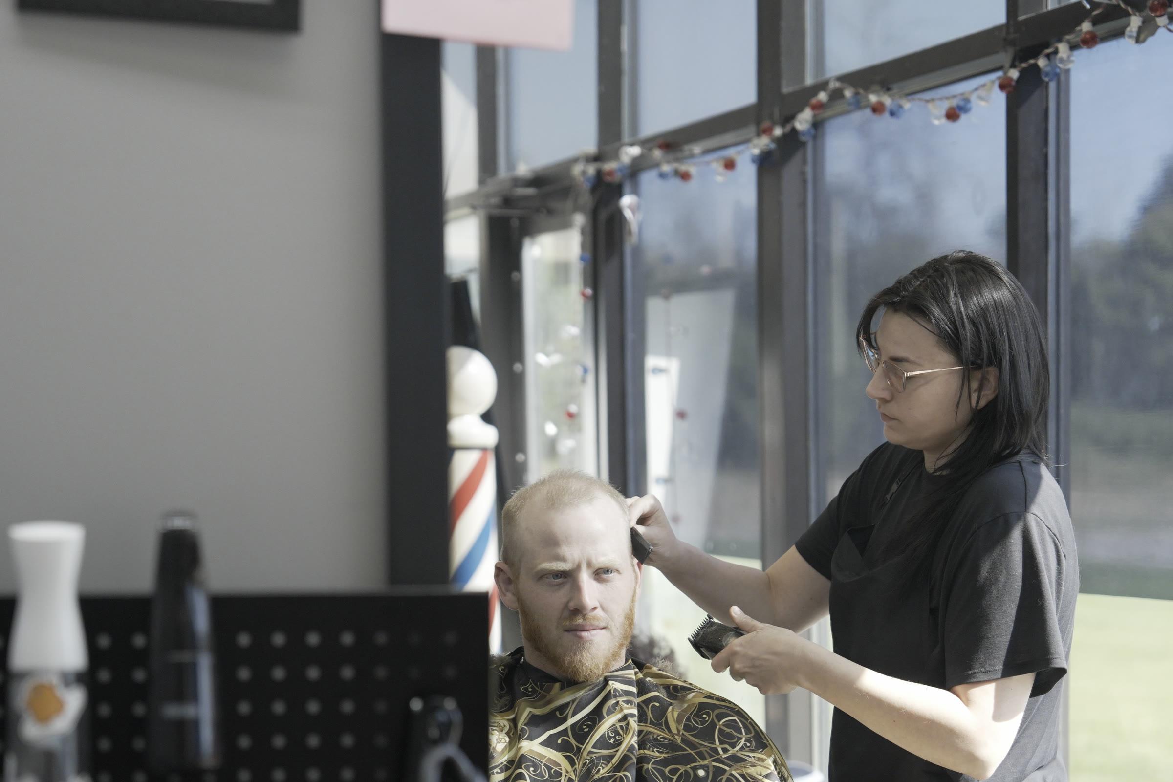 A student cutting a client's hair, the pair reflected in the station mirror with a barber pole visible through the window behind.
