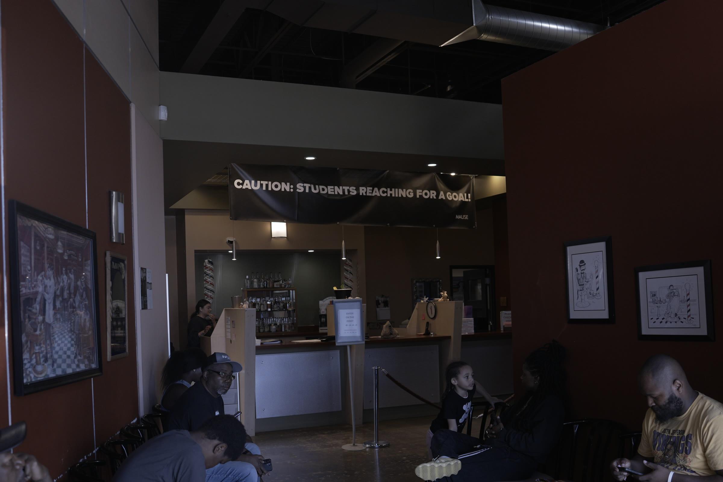 The waiting area inside the front door, clients seated along the wall and the reception counter beyond, under a banner reading Caution: students reaching for a goal.