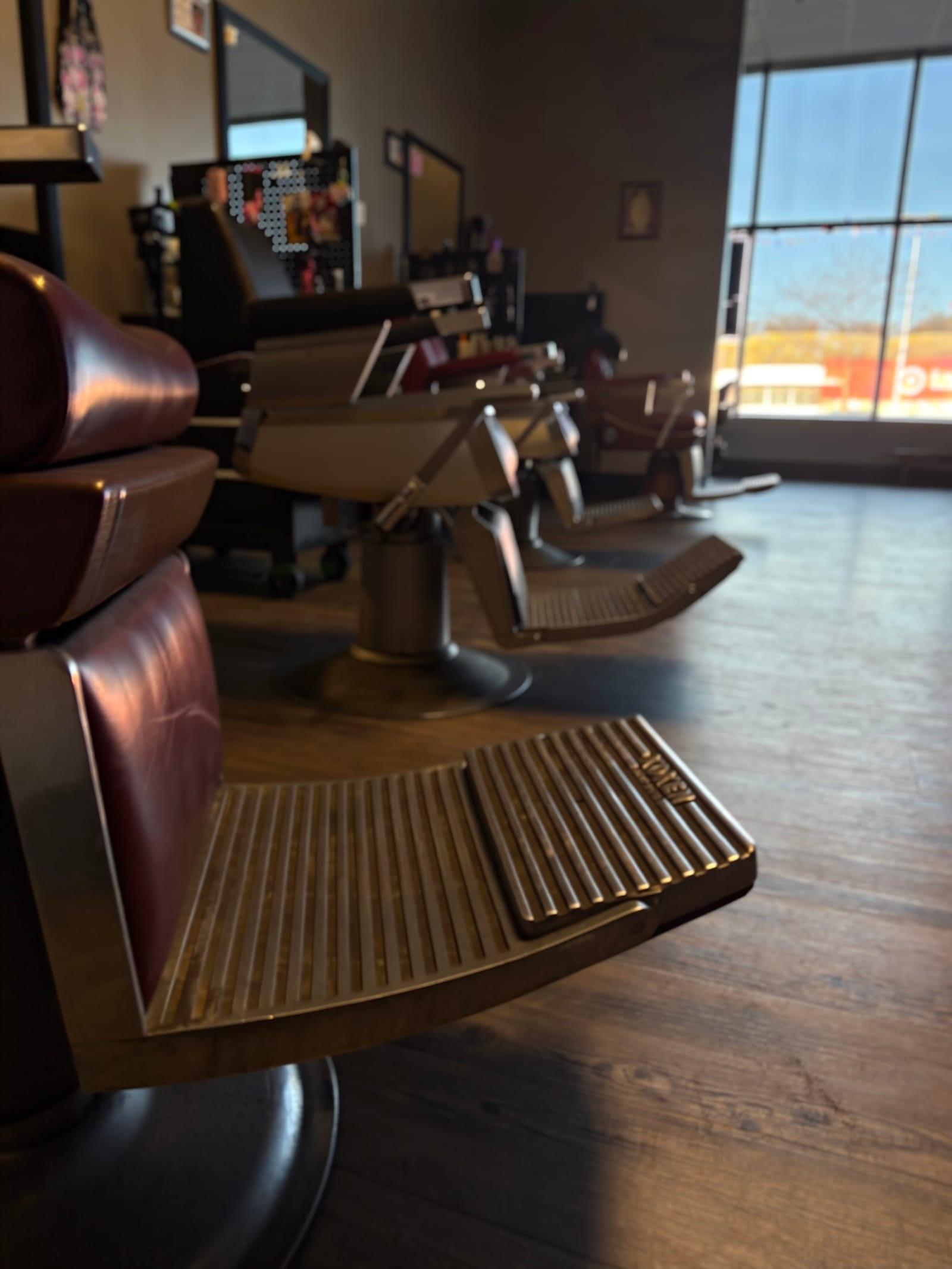 A row of barber chairs on the student floor, seen low from beside the footrest of the nearest chair, the front windows bright at the far end.